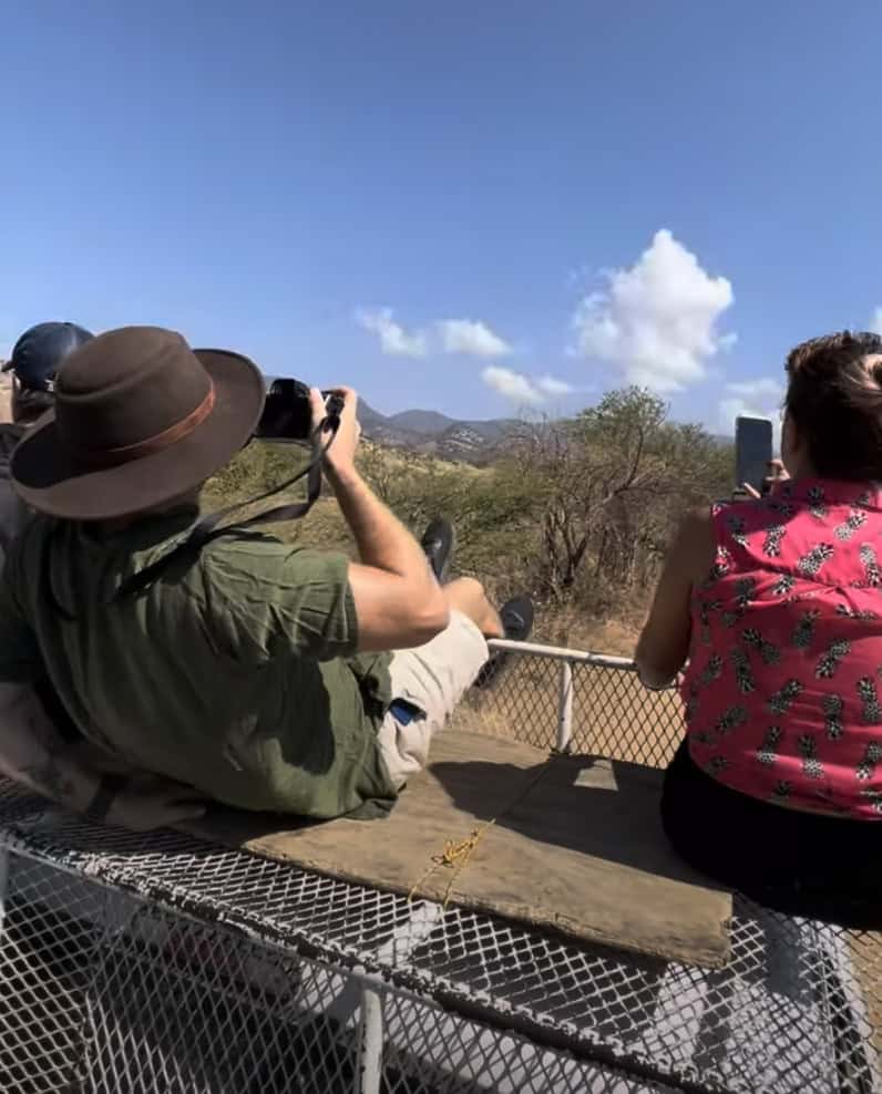 Two people on a safari-style truck snap photos of the desert landscape under a clear blue sky with puffy clouds.
