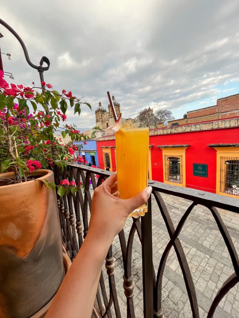 A hand holding a tall orange cocktail over a charming balcony view of colorful colonial buildings and a cathedral in Oaxaca, Mexico.