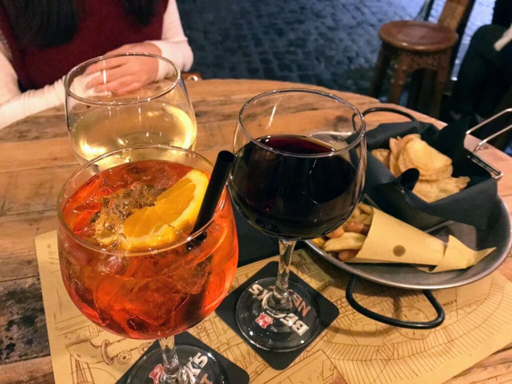 A wooden table at a cozy bar with three drinks: an Aperol spritz with an orange slice, a glass of red wine, and a glass of white wine, with a basket of potato chips and cheese in the background.