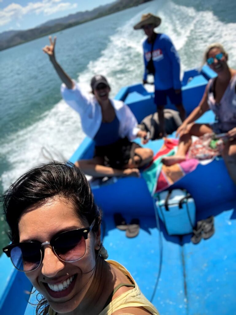 A group of friends enjoying a boat ride on a sunny day, with one woman smiling in the foreground for a selfie and others relaxing behind her on a bright blue boat.