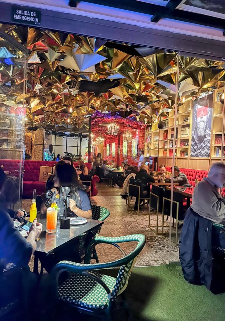 A lively restaurant scene with moody lighting, gold geometric ceiling decor, and guests dining in a chic and artistic setting.