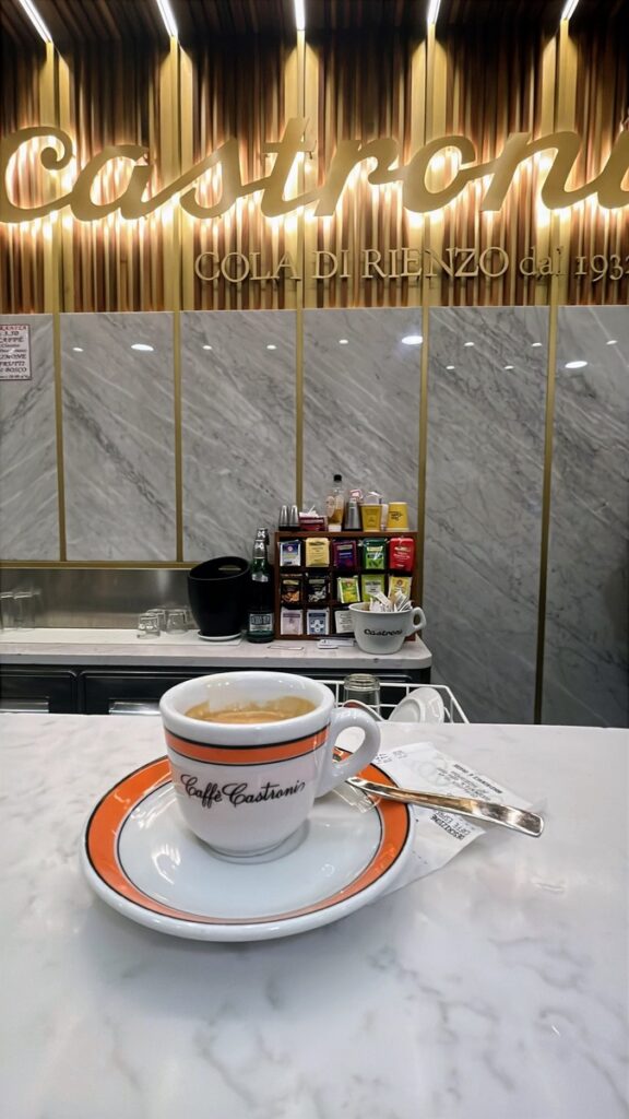 White espresso cup filled with coffee on a marble counter inside Caffè Castroni. The gold and marble café interior features a glowing “Castroni” sign and a display of colorful tea and coffee products in the background.