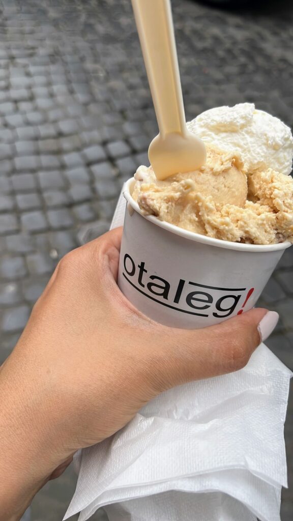 Hand holding a cup of creamy gelato topped with whipped cream on a cobblestone street. The white cup reads “Otaleg!” and a small plastic spoon is tucked into the scoop of gelato.