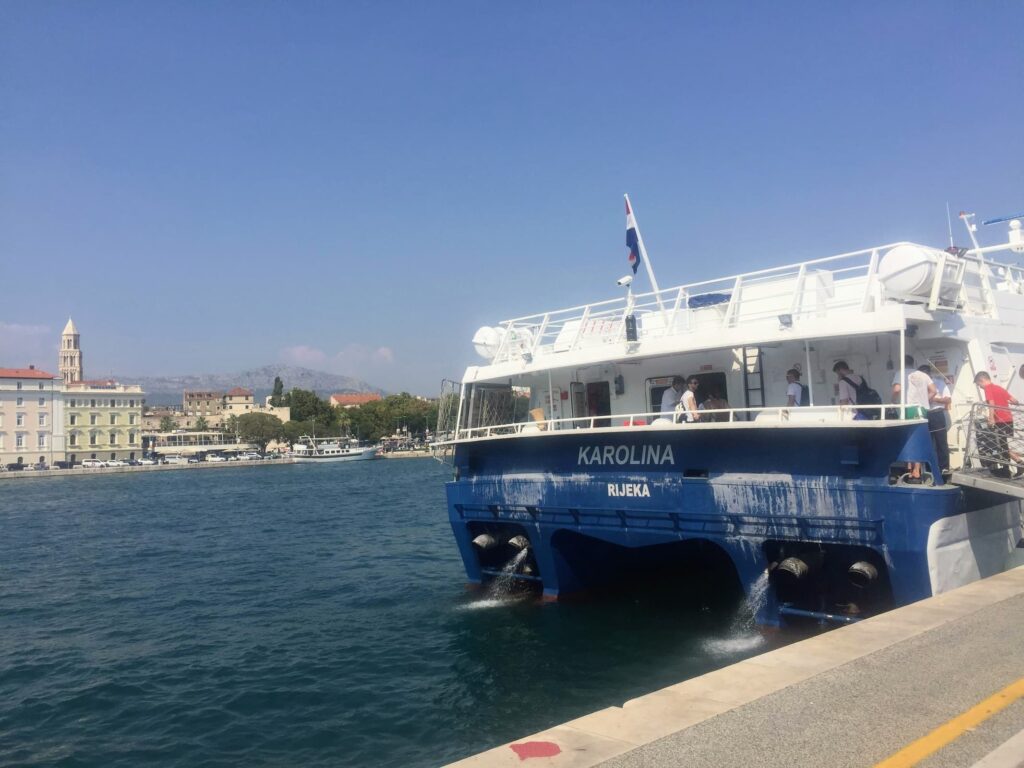 A blue and white passenger ferry named "Karolina" from Rijeka is docked at a waterfront in Split, Croatia. The city skyline, including a prominent bell tower and distant mountains, is visible under a bright blue sky.