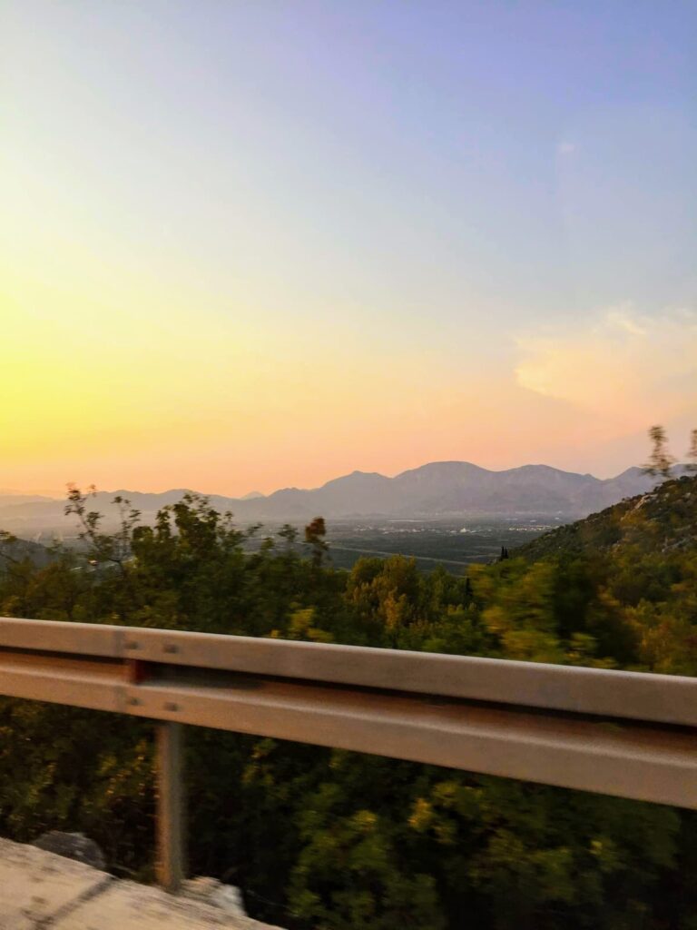A golden sunset casts a warm glow over rolling hills and distant mountains, as seen from a roadside with a metal guardrail. The lush greenery in the foreground contrasts with the pastel hues of the sky.