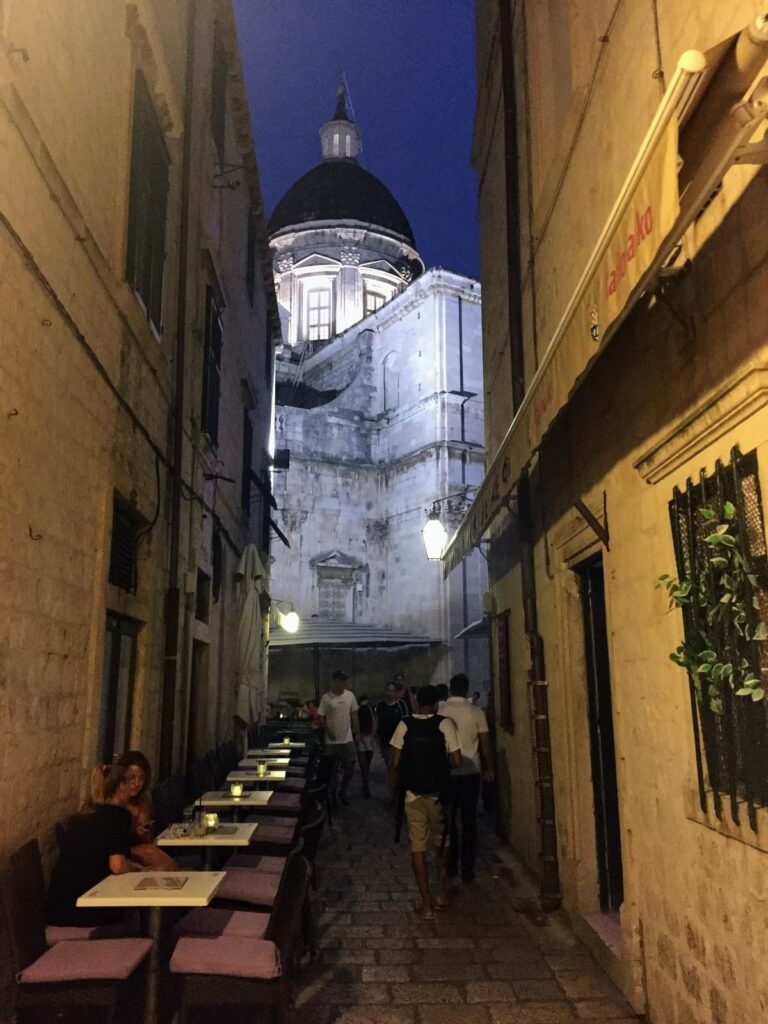 A dimly lit, narrow street in Dubrovnik, Croatia, leads to a grand domed historic building. Outdoor seating with people dining and walking along the cobblestone path creates a lively evening atmosphere.