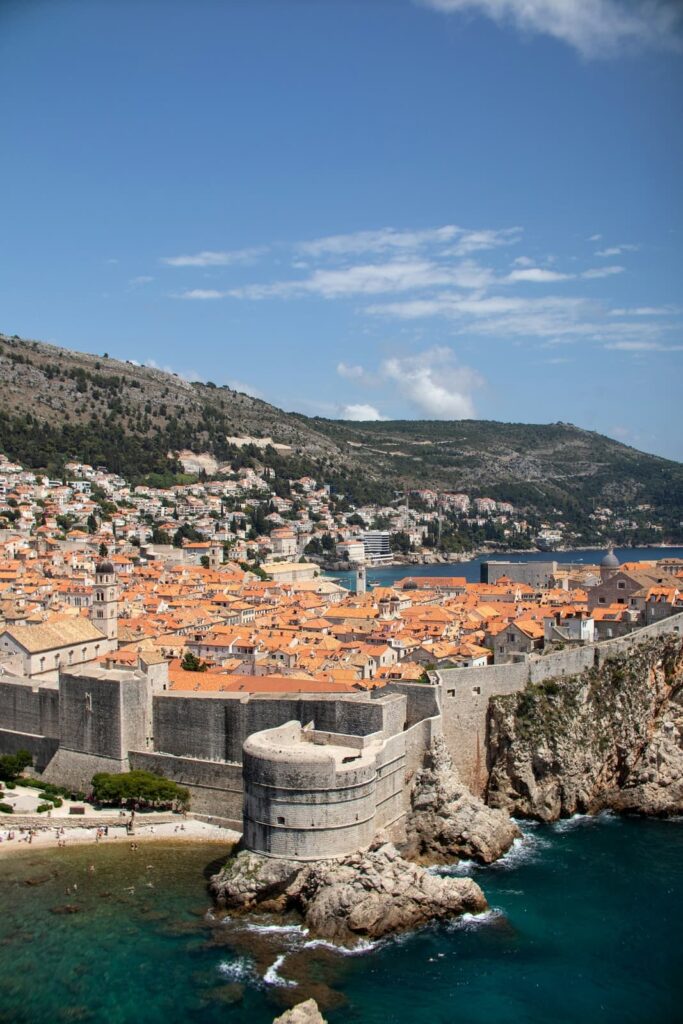 Dubrovnik’s historic Old Town, with its iconic terracotta rooftops, fortified stone walls, and blue Adriatic waters, is captured from an elevated perspective. The round fortress on the coastline stands prominently in the foreground.