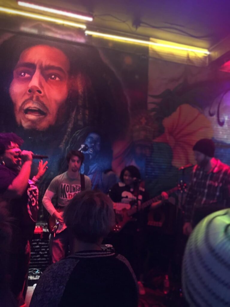 A dimly lit live music venue with a reggae band performing in front of a vibrant mural of Bob Marley. The crowd is watching as a singer and guitarist perform under colorful neon lights.