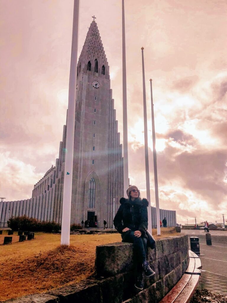 A person in a dark coat and knit hat sits on a stone wall in front of Hallgrímskirkja, the iconic Lutheran church in Reykjavík, Iceland in October. The tall, tiered concrete facade rises dramatically into a glowing cloudy sky, flanked by bare flagpoles.
