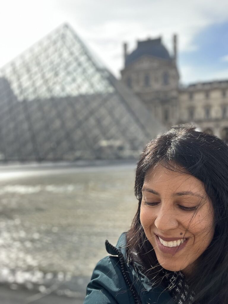 A woman smiles with her eyes closed, standing in front of the Louvre’s iconic glass pyramid on a bright, breezy day, with the historic museum building in the background.