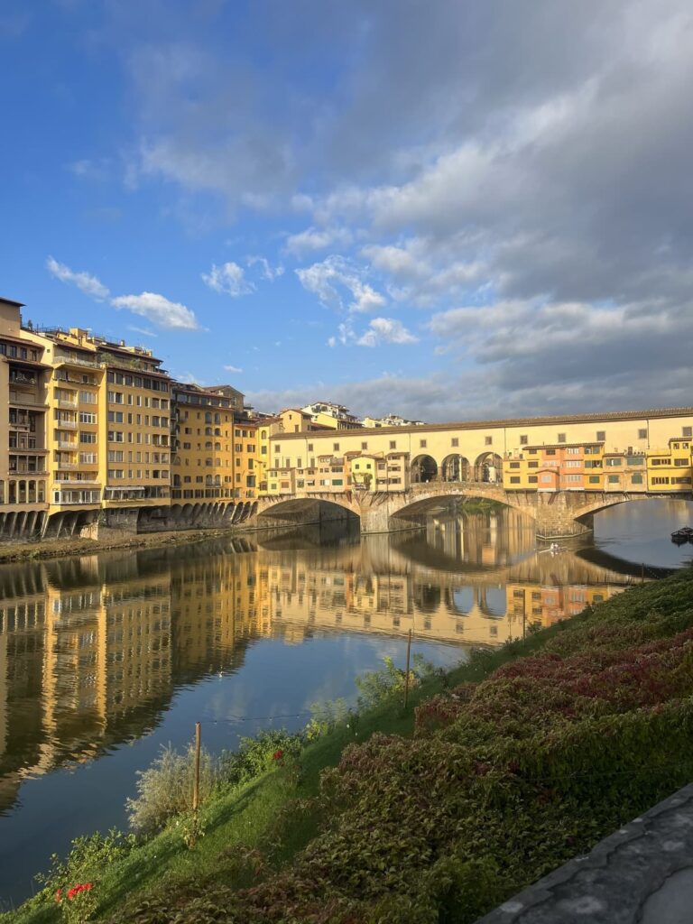 A scenic shot of the Ponte Vecchio bridge in Florence, with golden buildings reflecting in the calm waters of the Arno River under a bright sky with puffy clouds.