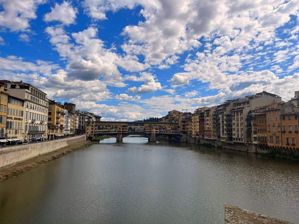 A wide shot of the Ponte Vecchio bridge crossing the Arno River, framed by old Florentine buildings on both riverbanks and a sky full of textured clouds.