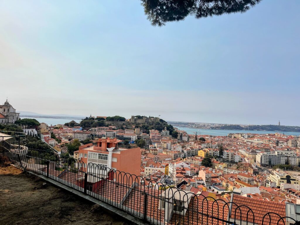 A panoramic view of Lisbon, Portugal, from a high vantage point. The cityscape features terracotta rooftops, colorful buildings, and the Tagus River in the background, with São Jorge Castle perched on a hill.