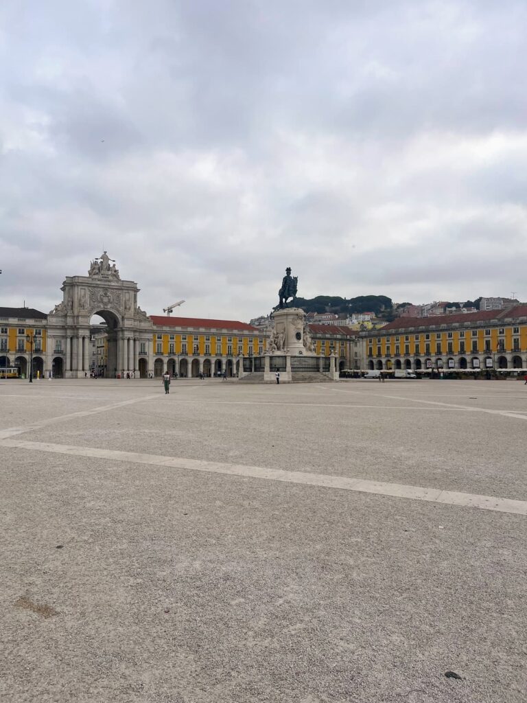A grand open square in Lisbon, surrounded by yellow buildings with arcades. The statue of King José I stands in the center, and the triumphal Rua Augusta Arch is visible in the background under an overcast sky.