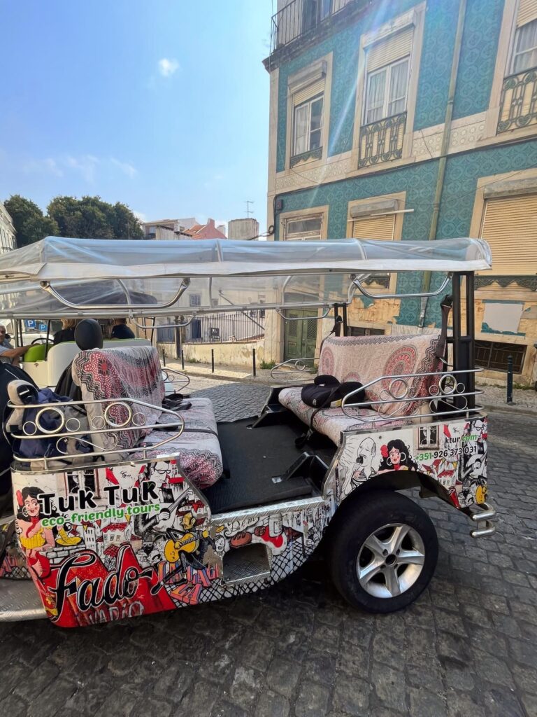 A vibrantly decorated tuk-tuk in Lisbon, covered in graffiti-style artwork featuring Fado music themes and city landmarks. The open-air vehicle is parked on a cobbled street next to a blue-tiled building.