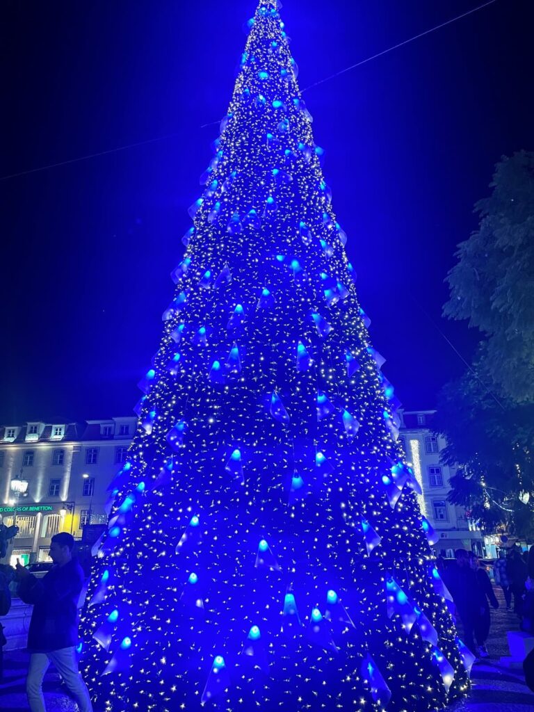 A towering Christmas tree is lit up with glowing blue and white lights in a public square at night, creating a dramatic holiday display. People gather around the base, snapping photos and soaking in the festive vibes.