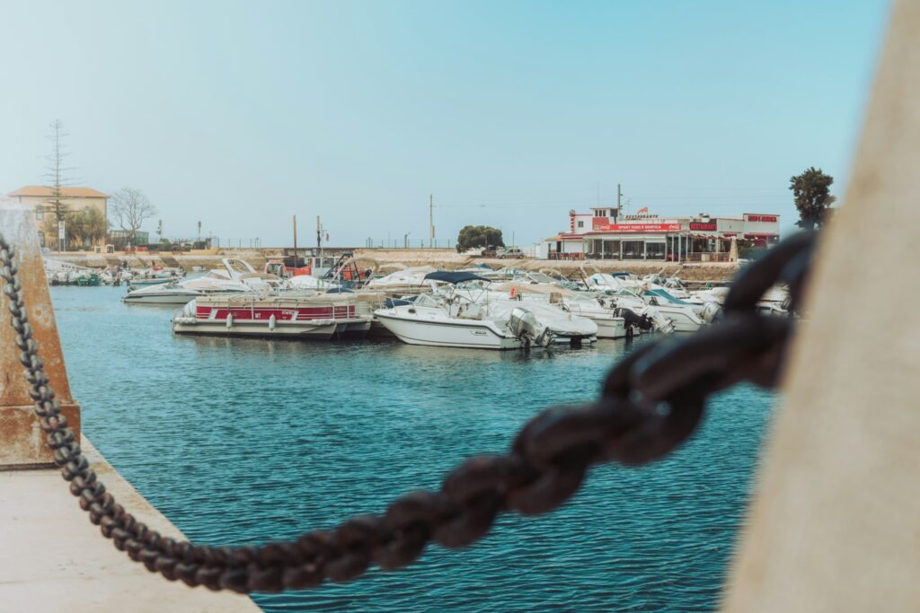 A peaceful marina filled with docked white boats and calm turquoise water, framed by a thick black chain in the foreground. A small building labeled "Restaurant" sits near the horizon under a clear blue sky.