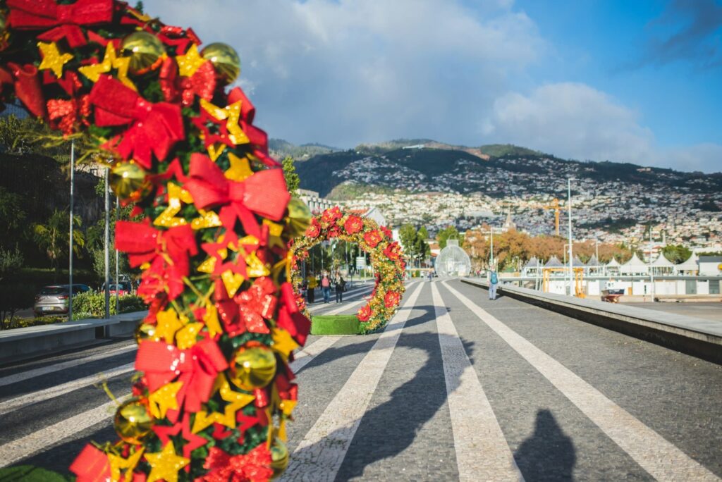 A sunlit promenade decorated with large holiday arches made of red bows, gold ornaments, and glittery yellow stars. In the background, a hillside town spreads across the mountains under a mix of clouds and blue sky, adding a scenic touch to the cheerful holiday vibe.