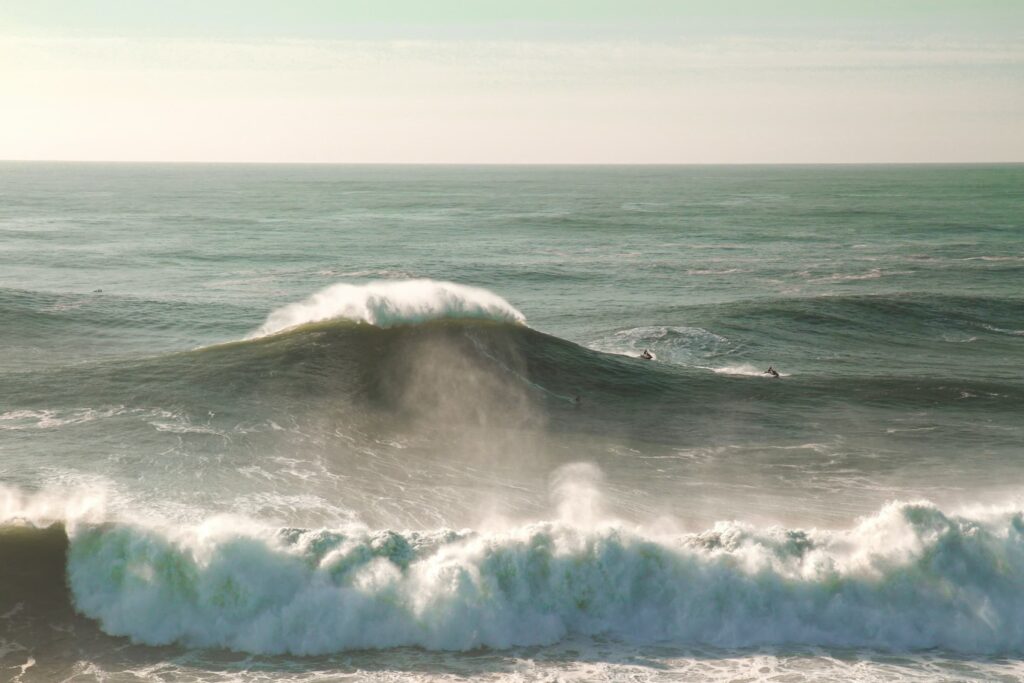 Massive ocean waves crash and curl under a hazy sky as a few surfers on jet skis ride the powerful swells. Mist rises off the surf, giving the scene an epic, high-energy feel.