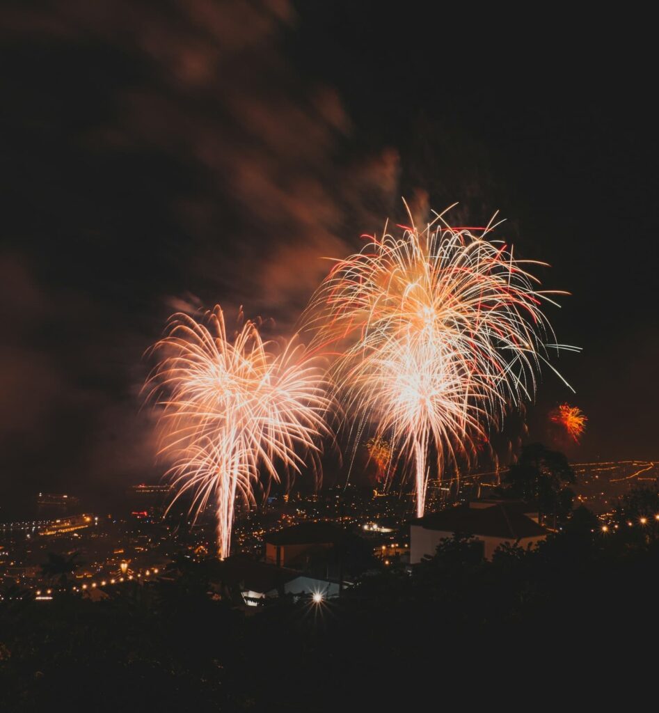 Bright, golden fireworks burst dramatically over a city at night, lighting up rooftops and casting a festive glow. Multiple fireworks explode in sync, illuminating the cloudy sky with vivid light trails.