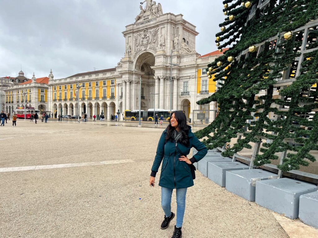 : A woman in a teal coat stands smiling near a modern metal Christmas tree structure in Lisbon’s Praça do Comércio. Behind her is the iconic Arco da Rua Augusta and the vibrant yellow façades of the surrounding buildings.
