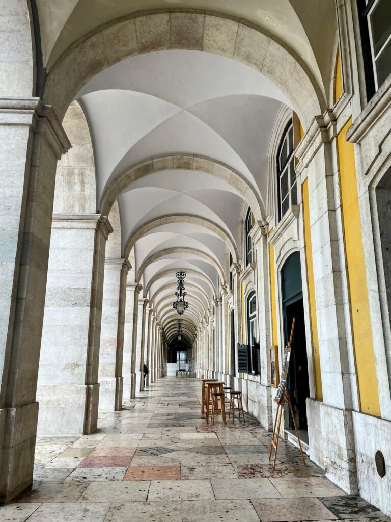 A long, elegant marble corridor in Lisbon with high vaulted ceilings and archways stretching into the distance. A few stools and easels line the side near a cozy café entrance.