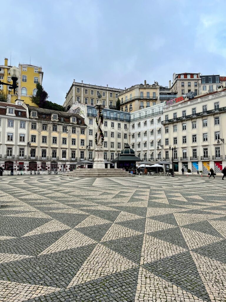 Rossio Square in Lisbon features intricate black-and-white patterned cobblestone pavement leading to a central columned statue. Classic Portuguese architecture and hills with clustered buildings frame the scene.