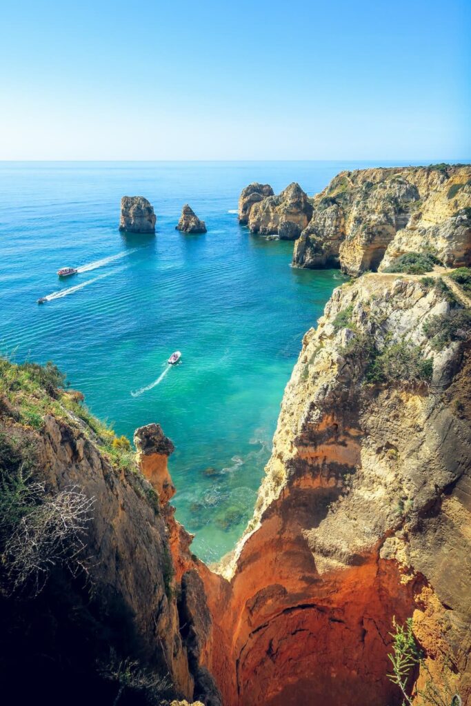 Clear turquoise waters surround the golden sea cliffs of Ponta da Piedade in Lagos, Portugal. Small boats leave soft wakes as they cruise through the natural rock formations jutting out of the ocean.