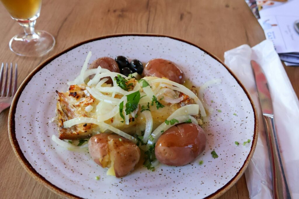 A traditional Portuguese plate of Bacalhau à Brás featuring salted cod, potatoes, black olives, onions, and herbs, garnished with fresh parsley. The dish is served on a rustic ceramic plate next to a cold drink and silverware.