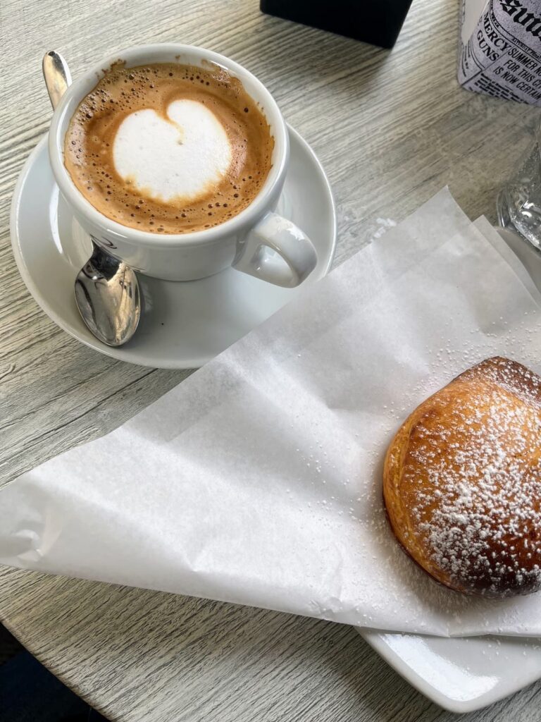 A cup of cappuccino with a heart-shaped foam design sits on a white saucer, accompanied by a spoon. Beside it, a golden-brown pastry dusted with powdered sugar rests on parchment paper atop a plate. The breakfast is set on a rustic wooden table, with a glass of water and a newspaper-themed napkin holder in the background.