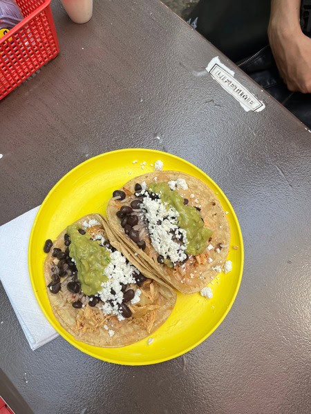 yellow plate with two tacos on it topped with cheese, black beans and guacamole on corn tortilla