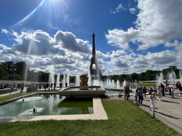View of Eiffel Tower, with green grass, fountains and people taking photos in front of it on a sunny day with clear blue skies