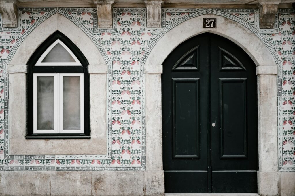 Ornate doorway of a Lisbon building featuring traditional Portuguese azulejos with a floral pattern. The black, arched double door contrasts with the white-framed window beside it.