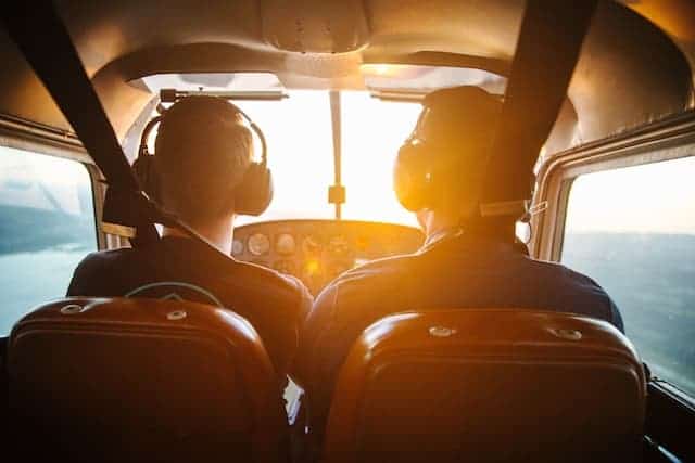 backs of two copilots in a small plane looking out ahead of window as they fly, sun streaming in