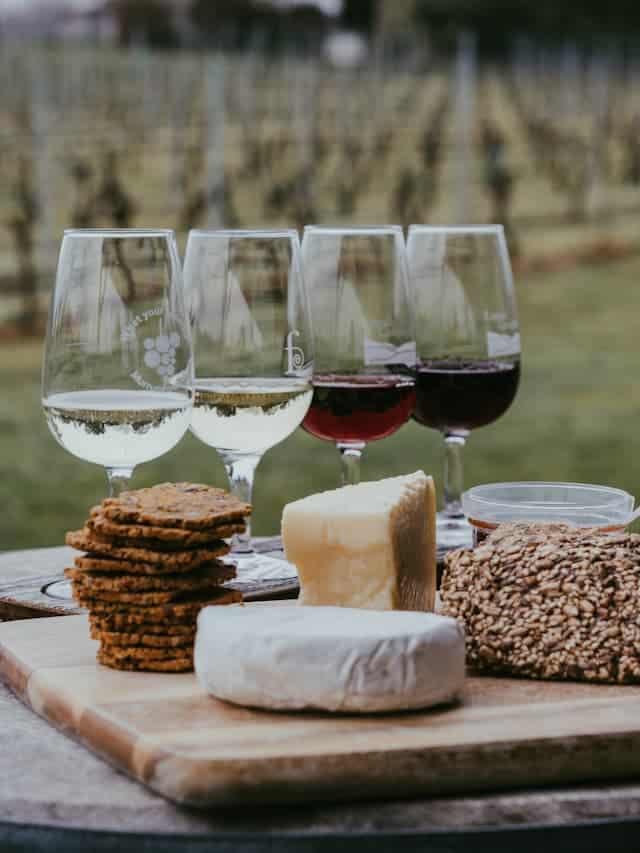 barrel with 2 red and 2 white wine glasses with cheese and crackers in front of a vineyard