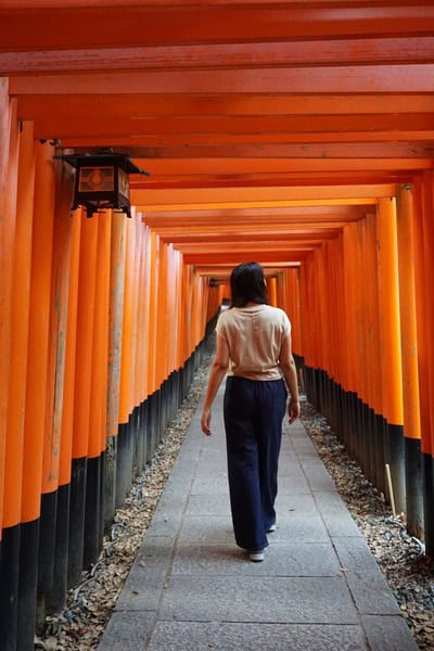 woman in dark jeans and a tan short0sleeved shirt walking under traditional, rectangular orange shrine structures in Japan