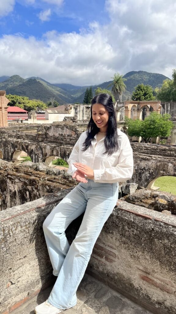 Woman in light wash blue jeans and a white, long sleeve collared linen shirt looks down at her hands, smiling, with mountains and ruins behind her in Antigua, Guatemala