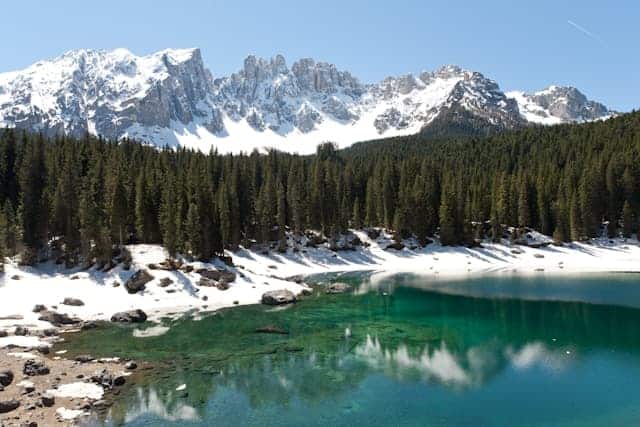 turquoise lake in Italy surrounded by snowy banks, thin trees and snowcapped mountains in the back