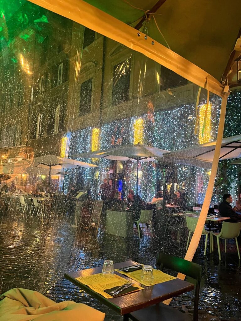 View of an outdoor café covered with transparent plastic sheets under yellow canopies during a rainy evening. Sparkling lights and cozy ambiance can be seen from the restaurant and street reflections on the wet pavement.