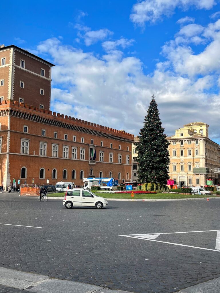 Piazza featuring a large decorated Christmas tree surrounded by historical buildings under a bright blue sky. Cars and people are seen in the square, emphasizing the lively yet calm atmosphere.