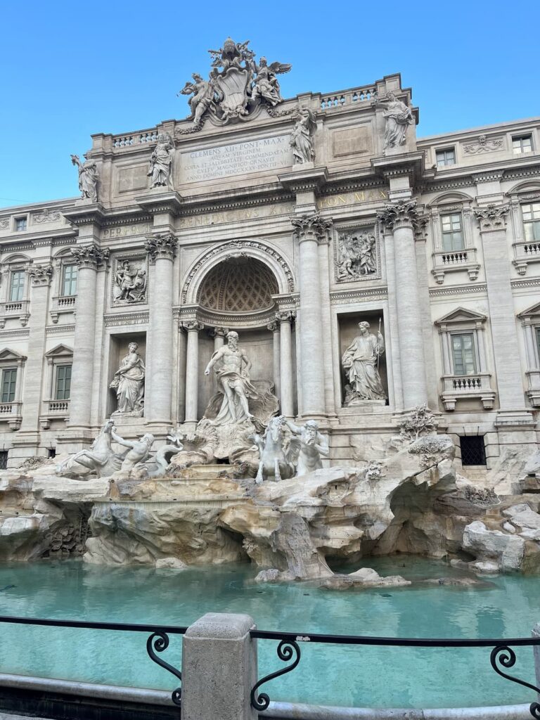 The grand Trevi Fountain with intricate marble sculptures of mythological figures and horses surrounded by cascading water into the clear turquoise pool below. The majestic architecture stands tall under the bright blue sky.