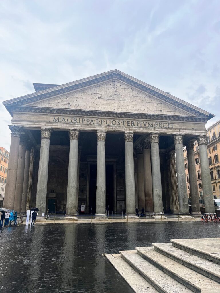 The majestic Pantheon with its grand portico and tall Corinthian columns standing on a wet, rainy square. Visitors with umbrellas are seen approaching the entrance under the overcast sky.