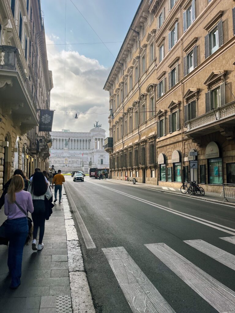 people walking on a sidewalk in Rome alongside a main street with buildings lining both sides of the street, and wedding cake building visible in the distance