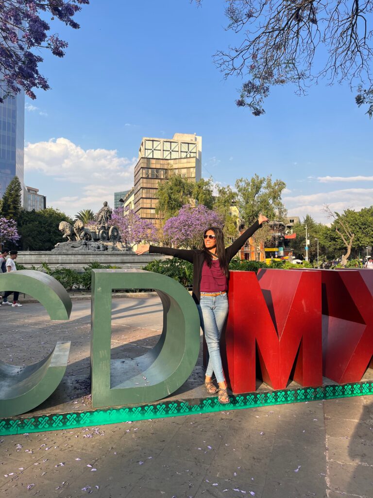 woman in jeans, black sweater and pink blouse poses with CDMX block letters in Mexico City