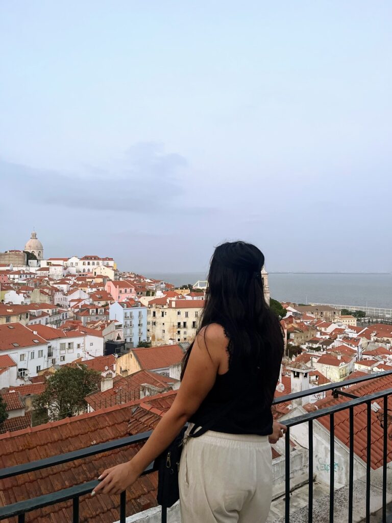 woman wearing travel capsule wardrobe pieces of cream linen pants and black sleeveless blouse looking out over the city of Lisbon. Her back is to the camera