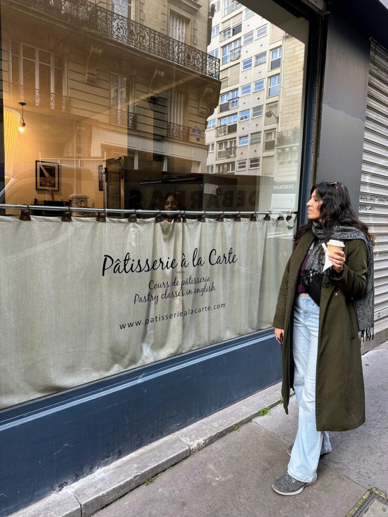 woman in light jeans and long green wool coat, unbuttened, holding a coffee cup standing in front of a bakery in Paris 