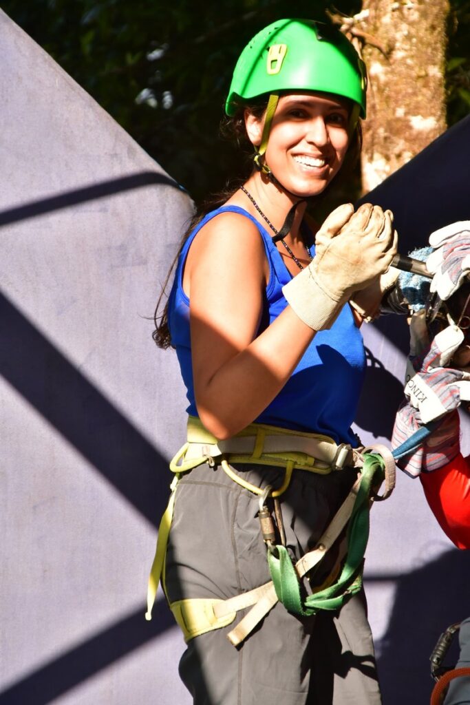 woman in ziplining gear including harness and helmet, wearing a blue workout tank top and grey flowy hiking pants
