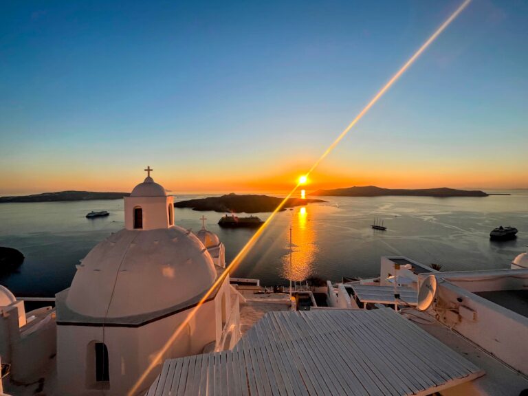 Orange sun setting over the sea with rays of light coming from it on two sides, diaganolly. A white stone dome in the foreground with a cross at the top.