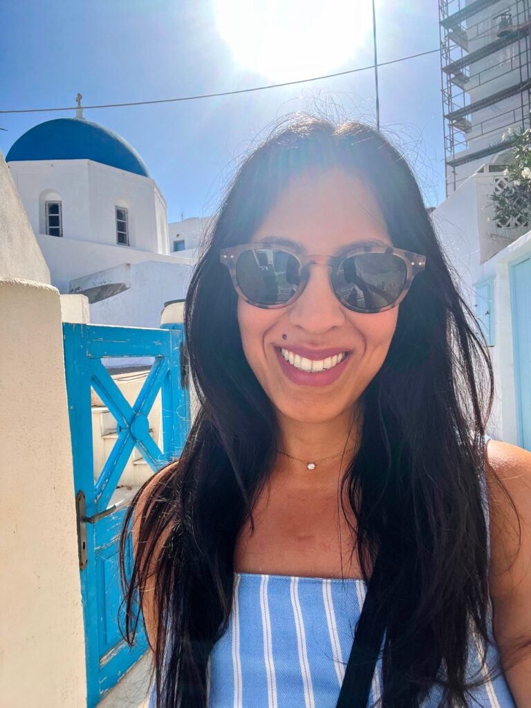 woman in blue and white striped dress, wearing sunglasses, smiling at camera with blue gate, whitewashed building and blue dome behind her in Santorini in October