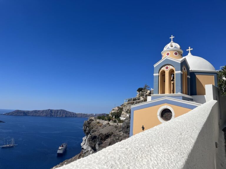 side view of mustard yellow colored church with blue lining and accents on a cliff next to the sea in Santorini Greece